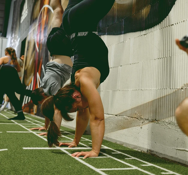 Sportlerin macht eine Handstand-Übung auf Kunstrasen in der Halle, während andere im Hintergrund trainieren.