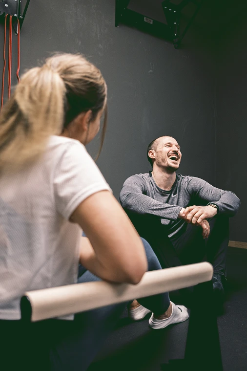 : Zwei Personen machen Trainingspause in der Halle; im Vordergrund sitzt eine Frau, im Hintergrund lächelt Coach Flex auf dem Boden.